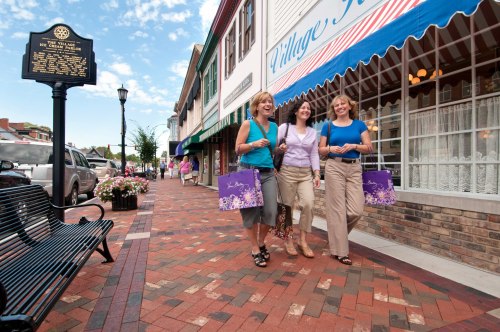 Three women walk together on a brick sidewalk, carrying shopping bags near a quaint storefront on a sunny day.