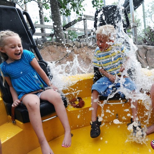 Two children on a water ride, splashing water and laughing, surrounded by trees and rocky scenery.