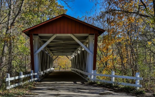 A red covered bridge surrounded by trees with autumn foliage, leading into a sunny path.