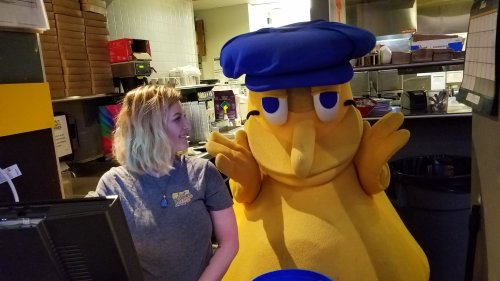 A person smiles next to a large, friendly yellow mascot wearing a blue hat in a restaurant setting.