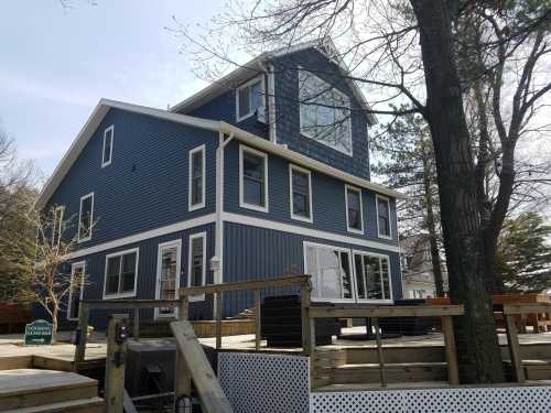 A large blue house with multiple windows, surrounded by a wooden deck and trees on a sunny day.