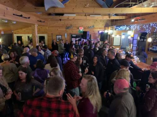 A lively crowd socializes in a rustic bar with wooden decor, drinks in hand, and festive lighting.