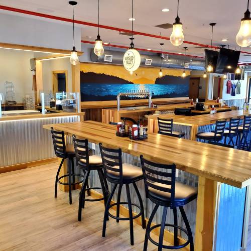 A modern bar with wooden tables and black chairs, featuring a scenic mural and pendant lights in a bright space.