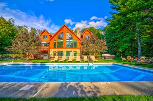A large wooden house with a pool and lounge chairs, surrounded by trees and a clear blue sky.