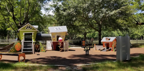 Colorful playground with small houses, slides, and play equipment surrounded by trees and grass.