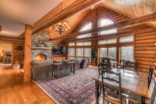 Cozy log cabin interior with a stone fireplace, large windows, and wooden beams, featuring a dining area and comfortable seating.