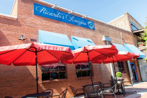 Exterior of Ricardo's Kouzzina with red umbrellas over outdoor seating and a blue sign on a brick building.