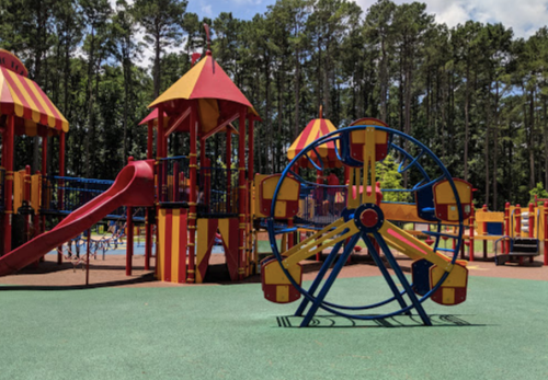 Colorful playground with a slide, climbing structures, and a Ferris wheel, surrounded by trees and blue sky.