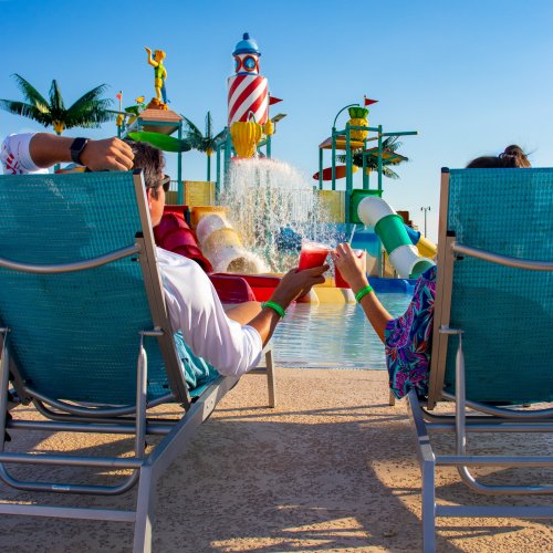 Two people relax in lounge chairs by a colorful water park, enjoying drinks with a fountain and palm trees in the background.