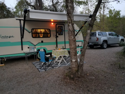 A vintage camper parked under trees, with a small table and chairs set up outside on a gravel path.