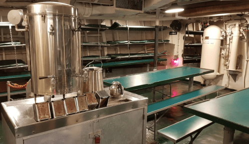 A ship's galley with stainless steel equipment, tables, and shelves, featuring a kettle and a water dispenser.