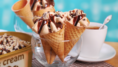 Three ice cream cones topped with chocolate drizzle and sprinkles, alongside a cup of dessert.
