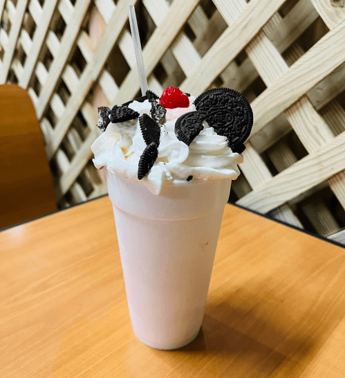 A tall milkshake topped with whipped cream, crushed Oreos, and a cherry, served in a clear cup on a wooden table.