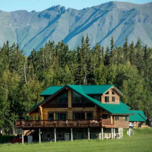 A log cabin with a green roof surrounded by trees and mountains in the background.