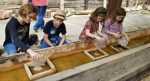 Four children are panning for gold in a shallow water trough, each using a bag to sift through the sand.