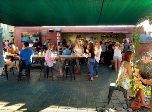 A lively outdoor bar scene with people socializing, seated at tables, and enjoying drinks under a shaded area.