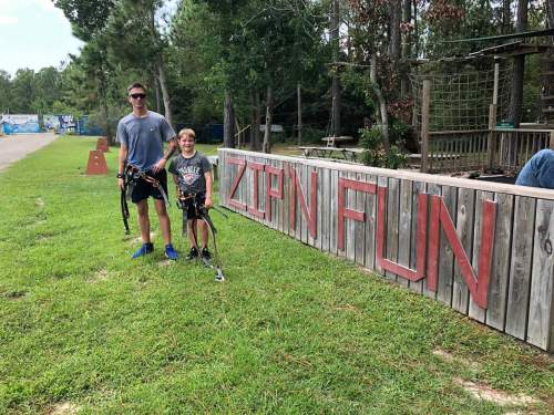 A man and a child stand in front of a wooden sign that reads "ZIPLINE FUN," both wearing harnesses for ziplining.