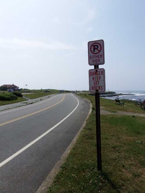 A winding road with a no parking sign and a warning sign about rocks, near a coastal area with grass and benches.