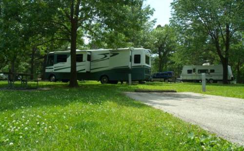 Two RVs parked in a grassy area surrounded by trees, with a paved path leading through the scene.