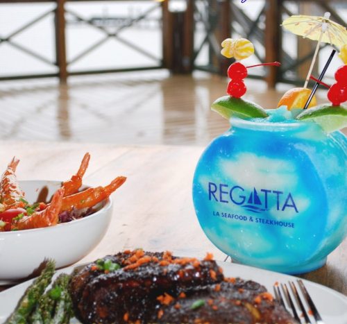A colorful cocktail in a fishbowl glass next to a plate of steak, shrimp, and asparagus on a wooden table.