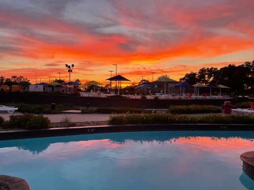 A vibrant sunset reflects in a pool, with umbrellas and a recreational area in the background.
