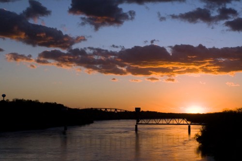 A serene sunset over a river, with clouds reflecting orange and purple hues, and a silhouette of a bridge in the foreground.