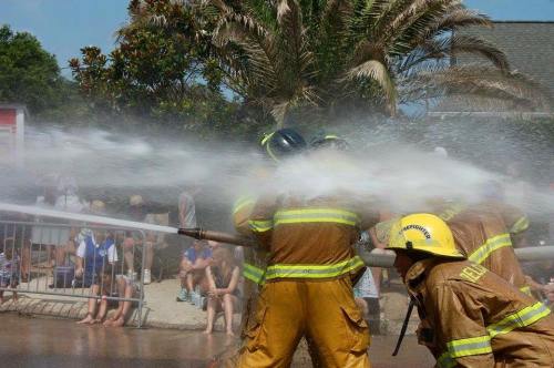 Firefighters in yellow gear operate a fire hose, spraying water in a lively outdoor setting with spectators in the background.
