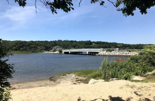 A serene river scene with a bridge in the background, surrounded by greenery and a sandy shore under a clear blue sky.