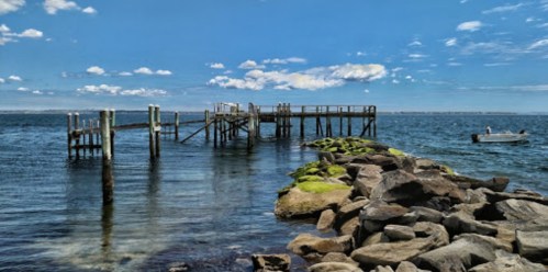 A wooden pier extends into calm waters, with a rocky shoreline and a small boat in the distance under a blue sky.