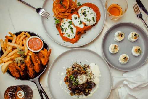 A table spread featuring fried chicken, pasta, a creamy dish with mushrooms, and sushi rolls, with drinks on the side.