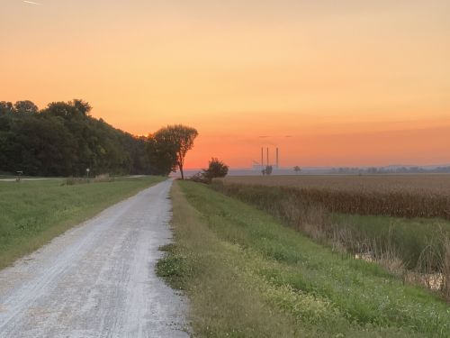 A gravel path lined by greenery leads to a sunset over fields, with power plant smokestacks in the distance.