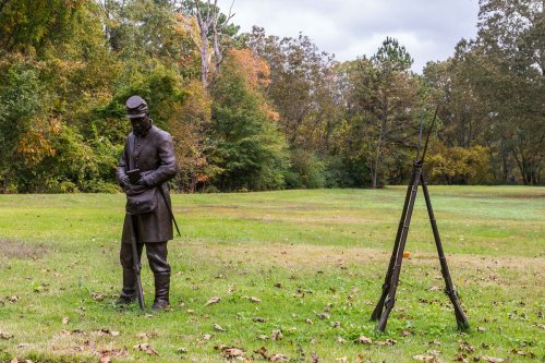 A bronze statue of a soldier stands in a grassy area, with trees and autumn foliage in the background.