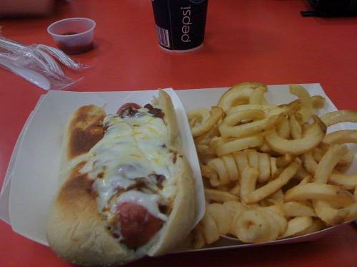 A hot dog topped with melted cheese and chili, served with a side of curly fries and a cup of soda.