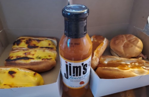 A bottle of Jim's famous sauce surrounded by hot dogs and a sandwich in a takeout box.