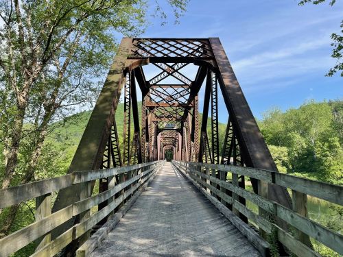 A metal bridge with a wooden walkway spans a lush green landscape under a clear blue sky.