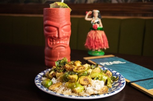 A plate of sautéed Brussels sprouts on rice, with a tiki mug and a hula dancer figurine in the background.