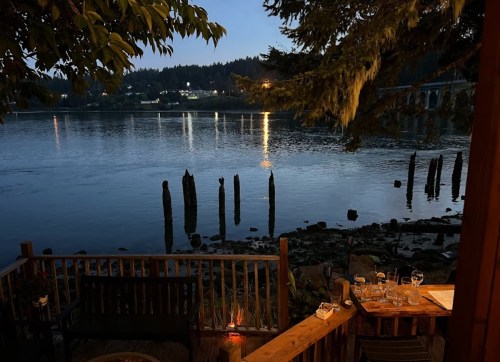 A serene riverside view at dusk, featuring water reflections, old wooden posts, and a cozy outdoor dining setup.