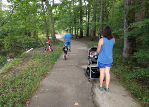 A family walks along a wooded path; a woman pushes a stroller, while two children ride bikes nearby.