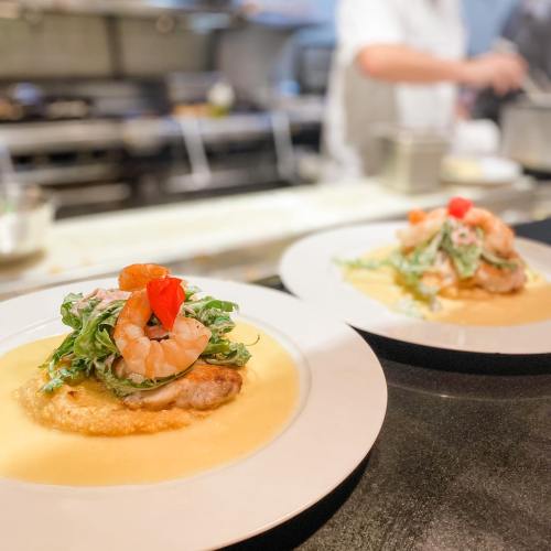 Two plates of shrimp atop a bed of greens and sauce, garnished with a flower, in a restaurant kitchen setting.