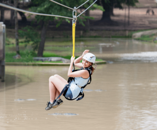 A person in a harness zip-lining over a muddy body of water, smiling and enjoying the adventure.