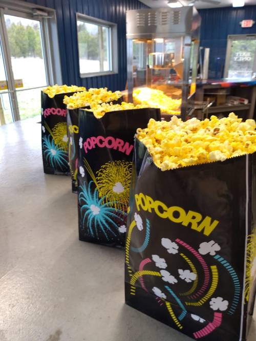 Colorful popcorn bags filled with popcorn sit on a counter near a popcorn machine in a bright, casual setting.