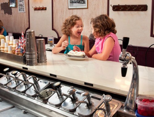 Two young girls excitedly share a dessert at a diner, sitting at a counter with ice cream supplies in the background.