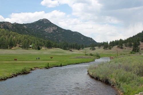 A serene landscape featuring a river, green fields, and horses grazing, with mountains in the background under a cloudy sky.