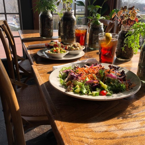 A wooden table set with a colorful salad, small bowls of sides, and drinks, surrounded by plants in jars.
