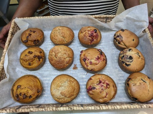 A person holds a basket of assorted muffins, including chocolate chip, blueberry, and berry varieties.