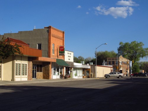 A quiet street scene featuring brick buildings, shops, and a clear blue sky with a few clouds.