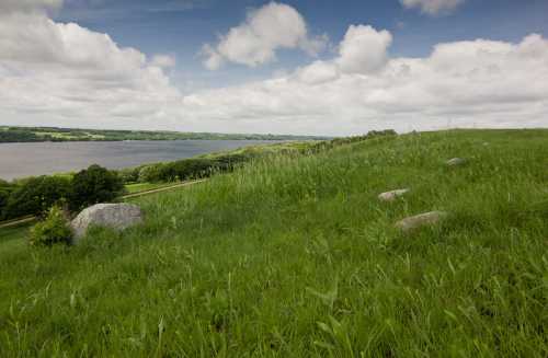 A grassy hillside with scattered rocks overlooks a calm river under a cloudy sky.