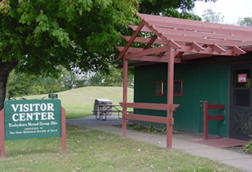 A small visitor center with a green exterior and red roof, surrounded by grass and trees, with a sign indicating its purpose.