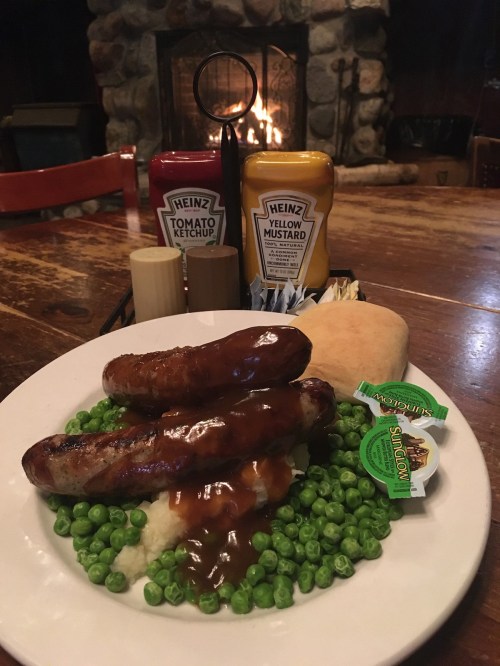 A plate with two sausages, green peas, a bun, and condiments, set on a rustic wooden table near a stone fireplace.