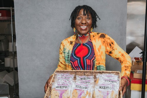 A smiling woman with dreadlocks holds a basket of Kizito products, wearing a colorful, patterned outfit.